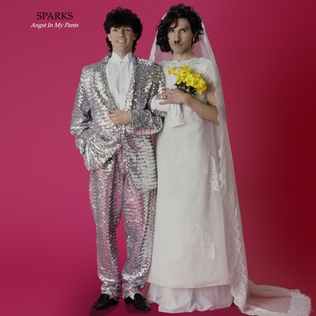Two men standing in front of a magenta background. The man on the left is wearing a natty silver suit. The man with the small moustache, on the right, is wearing a simple but beautiful wedding dress and carrying a small yellow bouquet.