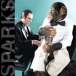 A man with in a black tuxedo with a small moustache sitting at a piano with a chimpanzee in a white tuxedo holding a vintage microphone in the foreground.