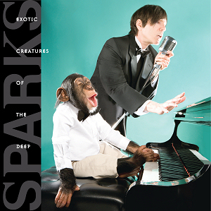 A chimpanzee in a white tuxedo sitting at a piano and seemingly singing. It does not have a small moustache. In the background a man in a black tuxedo sings into a vintage microphone.