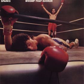 Two men in a boxing ring. In the foreground one is lying face down with one glowed hand on the ropes and the other hanging off the edge of the ring. The background the other is standing in a victory pose. He has a small moustache.
